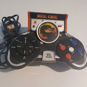 May include: A black and silver Mortal Kombat plug-and-play video game controller with a central dragon logo, a directional pad labeled with jump, duck, flip, and move, and action buttons for high punch, low punch, high kick, low kick, and block.