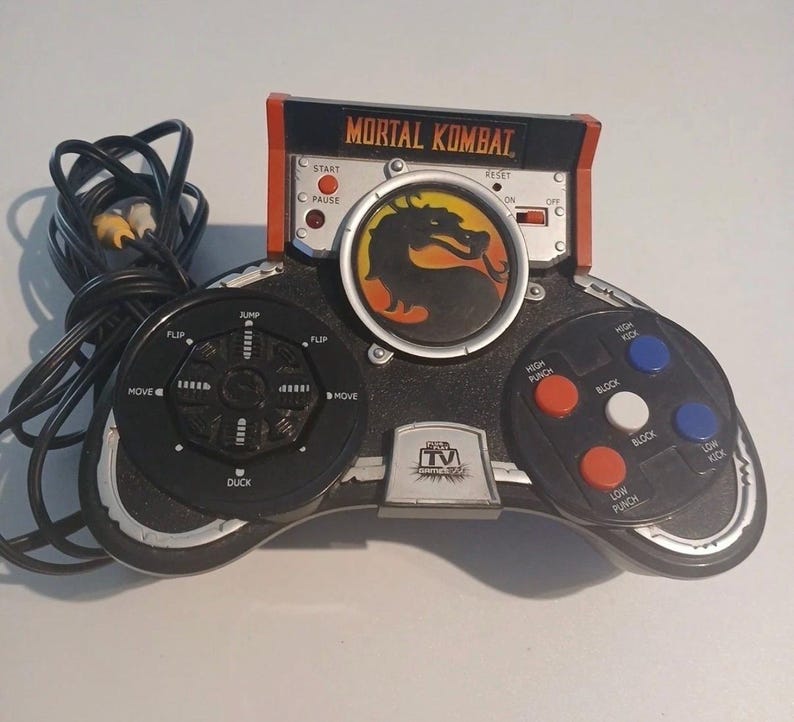 May include: A black and silver Mortal Kombat plug-and-play video game controller with a central dragon logo, a directional pad labeled with jump, duck, flip, and move, and action buttons for high punch, low punch, high kick, low kick, and block.