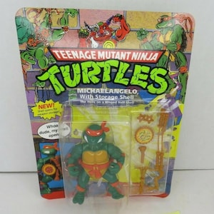 May include: A Teenage Mutant Ninja Turtles action figure of Michelangelo, with a hinged storage shell. The figure is green with an orange mask and a yellow shell. The packaging includes the text "NEW! Shell opens for weapon storage!"