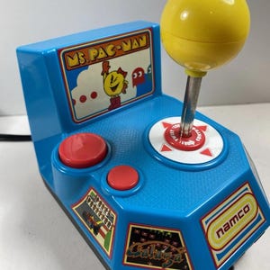 Puede incluir: Un controlador de juego arcade Ms. Pac-Man azul vintage con un joystick amarillo y botones rojos. El controlador presenta un gráfico de Ms. Pac-Man y el logotipo de Namco. Un objeto de colección de juegos retro.