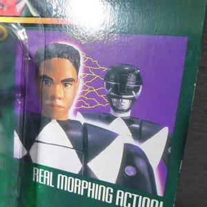 May include: A packaged action figure featuring a black and white Power Ranger with a helmet. The packaging includes a portrait of a person and the text "REAL MORPHING ACTION!" on a green background with purple accents.