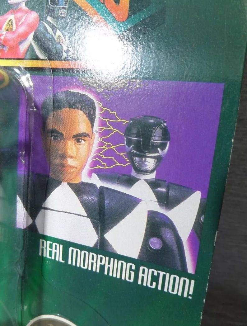 May include: A packaged action figure featuring a black and white Power Ranger with a helmet. The packaging includes a portrait of a person and the text "REAL MORPHING ACTION!" on a green background with purple accents.