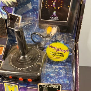 以下が含まれることがあります： ヴィンテージのAtariプラグアンドプレイゲームシステム。ジョイスティックと「Centipede」ゲームを表示する画面が特徴です。「Plug it in and play!」というテキストと、「Adventure」や「Yar's Revenge」などのゲームタイトルがパッケージに含まれています。