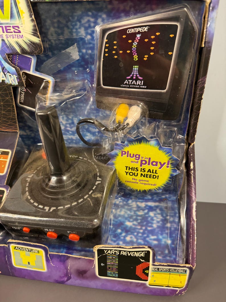 以下が含まれることがあります： ヴィンテージのAtariプラグアンドプレイゲームシステム。ジョイスティックと「Centipede」ゲームを表示する画面が特徴です。「Plug it in and play!」というテキストと、「Adventure」や「Yar's Revenge」などのゲームタイトルがパッケージに含まれています。