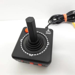 Op de afbeelding: Zwarte Atari joystick met een zwarte hendel en rode knoppen. De joystick heeft een cirkelvormig ontwerp met een witte stippellijn. Een zwarte kabel met een oranje band en een gele connector is bevestigd.