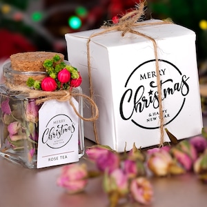 May include: A clear glass jar filled with dried rose petals, topped with a cork and decorative accents. A white gift box with "Merry Christmas & Happy New Year" text is tied with twine. Rose tea is the product.
