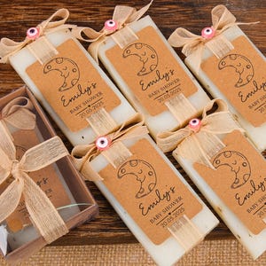 Może przedstawiać: Upominki na baby shower z prostokątnymi mydełkami i etykietami. Każde mydełko ma etykietę z napisem "Emily's BABY SHOWER 20.05.2025" i wzorem półksiężyca. Mydła są związane beżowymi wstążkami i ozdobami.