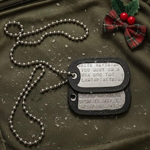 Puede incluir: Un par de placas de identificación de estilo militar con una cadena de bolas plateada. Las etiquetas son negras con letras plateadas que dicen "WRITE ANYTHING YOU WANT ON A USA DOG TAG". Un pequeño lazo a cuadros con bayas de acebo está en el fondo.