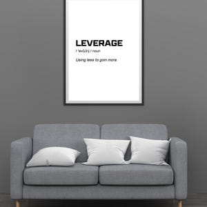 May include: A framed print with the word "LEVERAGE" in bold black font, along with its pronunciation and definition: "Using less to gain more." The print hangs above a gray sofa with white pillows.