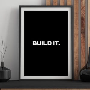 May include: Framed art print with a black background and the white text "BUILD IT." The artwork is in a black frame with a white border. The print is displayed on a black surface with decorative vases.