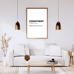 May include: A framed print with the word "COMMITMENT" in bold, black text. Below the word is the pronunciation and definition: "Staying even when it's hard." The print hangs above a white sofa in a modern living room.
