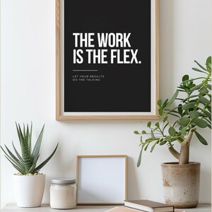 Könnte beinhalten: Ein rechteckiges Wandbild in einem hellen Holzrahmen mit schwarzem Hintergrund und weißem Text, der THE WORK IS THE FLEX lautet. Darunter steht in kleinerem weißem Text LET YOUR RESULTS DO THE TALKING, getrennt durch eine dünne weiße Linie.