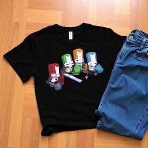 May include: Black t-shirt featuring a cartoon graphic of four knights with different colored helmets: red, green, orange, and blue. The knights are holding weapons and shields. A pair of blue jeans is next to the shirt on a wooden surface.