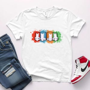 May include: White t-shirt with a graphic of four colorful characters wearing helmets. The characters are in a row and have different colored backgrounds.
