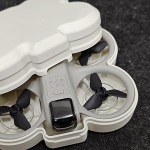 May include: A small, gray drone with four black propellers, housed in a white, cloud-shaped protective case. The drone has a black camera and control buttons. The case is open, revealing the drone's components.