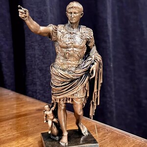 Könnte beinhalten: Bronzestatue eines römischen Kaisers, der mit dem rechten Arm zeigt. Die Figur trägt eine detaillierte Rüstung und eine drapierte Toga. Eine kleine Figur befindet sich zu Füßen des Kaisers. Die Statue ist auf einem rechteckigen Sockel montiert.
