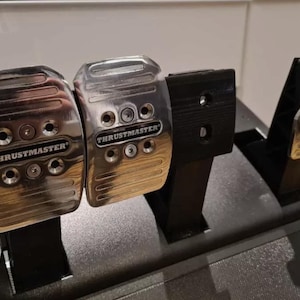 May include: A set of silver and black Thrustmaster racing pedals. The pedals are made of metal and have the Thrustmaster logo. The set includes an accelerator, brake, and clutch pedal, mounted on a black base.
