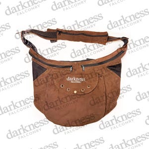 May include: A brown suede shoulder bag with multiple zippered compartments and a flap pocket. The bag features the text "darkness FALCONRY" and has a wide, adjustable strap. The bag is designed for carrying items.