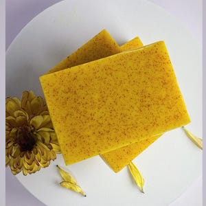 Può includere: Due saponette gialle con una texture punteggiata, disposte su un piatto bianco con petali di fiori gialli.
