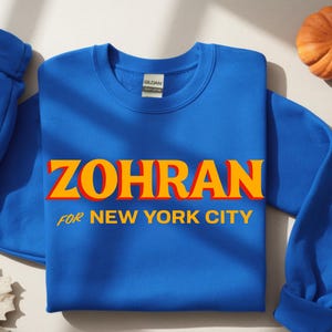 Może przedstawiać: Królewska niebieska bluza z napisem "ZOHRAN" w pomarańczowych i żółtych literach, z napisem "FOR NEW YORK CITY" poniżej. Na zdjęciu znajduje się również mała dynia i liść.