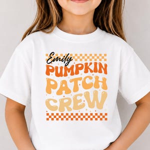 May include: A white short-sleeved t-shirt featuring a graphic design with orange and yellow wavy text that reads Emily PUMPKIN PATCH CREW, framed by orange and white checkered borders at the top and bottom, worn with light blue denim jeans.