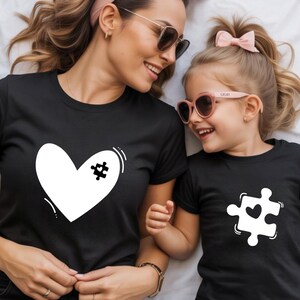 以下が含まれることがあります： 白のグラフィックが入った2枚の黒いTシャツ。1枚のシャツには、パズルのピースが付いた白いハートが、もう1枚にはハートが付いたパズルのピースが描かれています。シャツは母親と子供が着用しており、どちらもサングラスをかけて笑顔です。