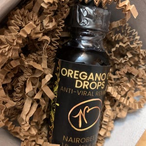 Puede incluir: Una pequeña botella de vidrio marrón oscuro con una etiqueta negra que dice "OREGANO OIL DROPS ANTI-VIRAL REMEDY" y un logotipo dorado con la letra "N". La botella está etiquetada como "NAIROBEES 1 fl oz (29.6 ml)".