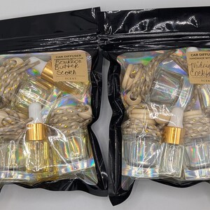 K&ouml;nnte beinhalten: Zwei schwarze Plastikt&uuml;ten mit Auto-Lufterfrischern. Eine T&uuml;te ist mit "Bourbon Butter Scotch" beschriftet, die andere mit "Midnight Cocktails". Jede T&uuml;te enth&auml;lt Glasflaschen, Holzperlen und goldfarbene Akzente.