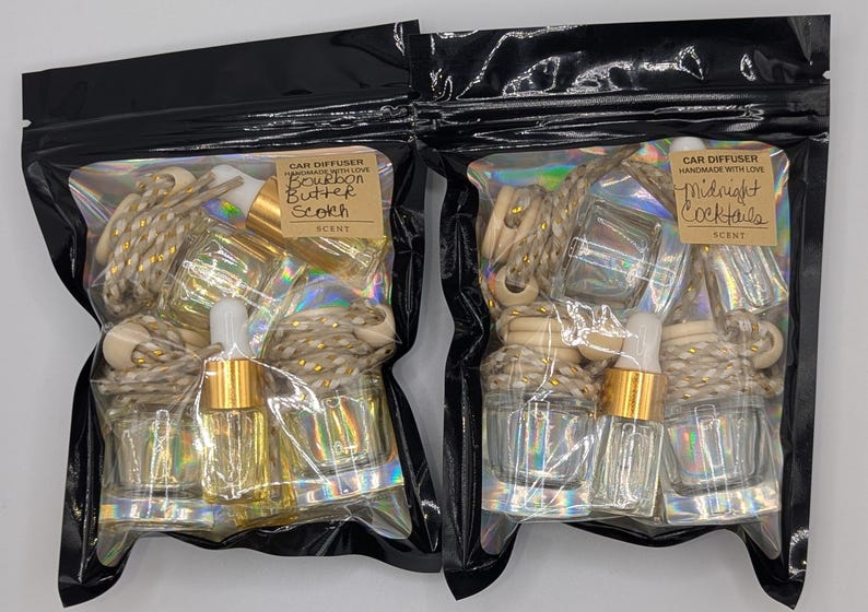 K&ouml;nnte beinhalten: Zwei schwarze Plastikt&uuml;ten mit Auto-Lufterfrischern. Eine T&uuml;te ist mit "Bourbon Butter Scotch" beschriftet, die andere mit "Midnight Cocktails". Jede T&uuml;te enth&auml;lt Glasflaschen, Holzperlen und goldfarbene Akzente.
