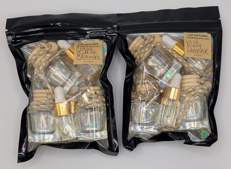 K&ouml;nnte beinhalten: Zwei schwarze Plastikt&uuml;ten gef&uuml;llt mit Lufterfrischern f&uuml;r Autos. Jede T&uuml;te enth&auml;lt mehrere kleine Glasflaschen mit goldfarbenen Deckeln und Holzperlen. Eine T&uuml;te ist mit "Cocoa Butter Cashmere" beschriftet, die andere mit "Wild Honey Lavender".
