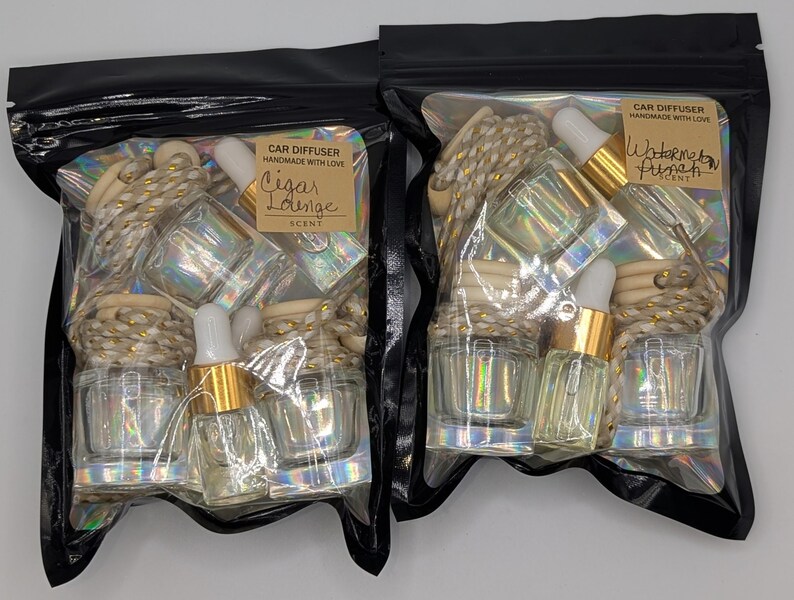 K&ouml;nnte beinhalten: Zwei schwarze Plastikt&uuml;ten mit Auto-Lufterfrischern. Jede T&uuml;te hat eine klare Glasflasche mit goldenem Verschluss und Pipette, zusammen mit einem Holzdiffusor und einem Etikett mit den D&uuml;ften "Cigar Lounge" und "Watermelon Punch".