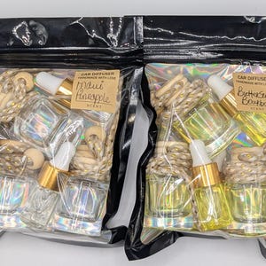 K&ouml;nnte beinhalten: Zwei Auto-Diffusor-Sets in transparenten Plastikt&uuml;ten. Eines ist mit "Maui Pineapple" und das andere mit "Butterscotch Bourbon" beschriftet. Jedes Set enth&auml;lt Glasflaschen mit goldenen Tropfern, Holzperlen und dekorativem Seil.