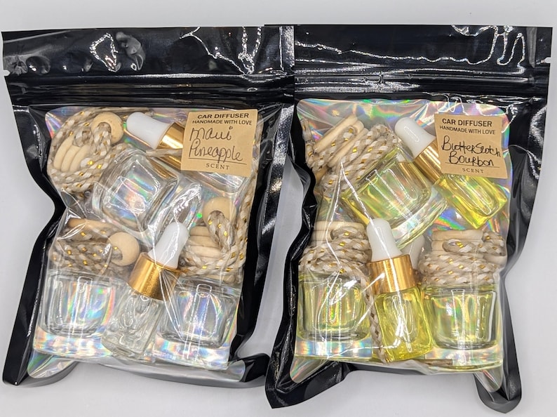 K&ouml;nnte beinhalten: Zwei Auto-Diffusor-Sets in transparenten Plastikt&uuml;ten. Eines ist mit "Maui Pineapple" und das andere mit "Butterscotch Bourbon" beschriftet. Jedes Set enth&auml;lt Glasflaschen mit goldenen Tropfern, Holzperlen und dekorativem Seil.
