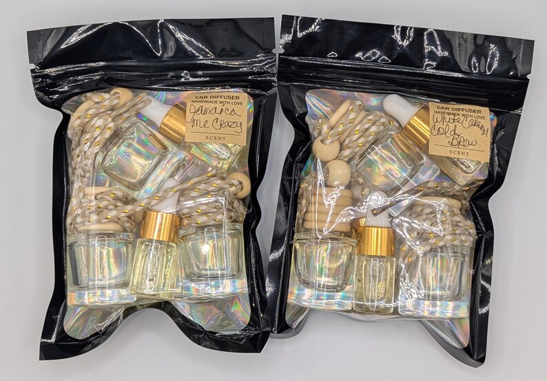 K&ouml;nnte beinhalten: Zwei schwarze Plastikt&uuml;ten mit Auto-Lufterfrischern. Jede T&uuml;te enth&auml;lt kleine Glasflaschen mit goldfarbenen Deckeln, Holzperlen und eine dekorative Schnur. Eine T&uuml;te tr&auml;gt die Aufschrift "Jamaica Me Crazy", die andere "White Candy Cold Brew".