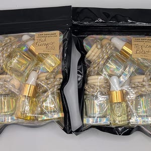 K&ouml;nnte beinhalten: Zwei schwarze Zip-Lock-Beutel mit Auto-Lufterfrischern. Jeder Beutel enth&auml;lt kleine Glasflaschen mit goldfarbenen Tropfern, Holzperlen und einem dekorativen Seil. Ein Beutel ist mit "Iced Cinnamon Rolls" und der andere mit "Tropical Blast" beschriftet.