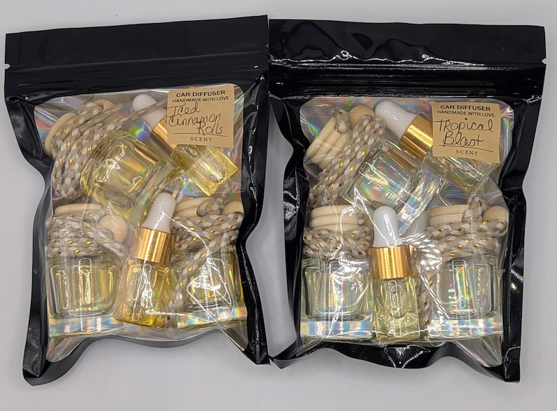 K&ouml;nnte beinhalten: Zwei schwarze Zip-Lock-Beutel mit Auto-Lufterfrischern. Jeder Beutel enth&auml;lt kleine Glasflaschen mit goldfarbenen Tropfern, Holzperlen und einem dekorativen Seil. Ein Beutel ist mit "Iced Cinnamon Rolls" und der andere mit "Tropical Blast" beschriftet.
