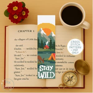 以下が含まれることがあります： 山と夕日のデザイン、テキスト「Stay Wild」、小さな小屋が描かれたクロスステッチのブックマーク。 ブックマークは開いた本の上にあり、コーヒーカップとコンパスが置かれています。