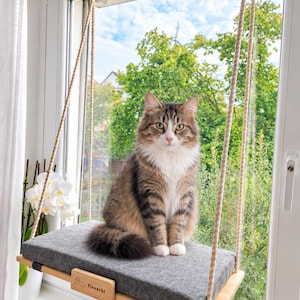 May include: A long-haired brown, white, and grey tabby cat sits on a rectangular grey felt cushion resting on a light-coloured wooden platform. The platform is suspended by thick beige ropes and features a small wooden tag labelled "Flauschi".