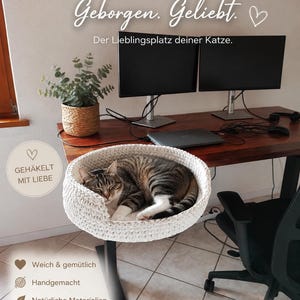 Peut inclure: Un panier pour chat rond et crocheté de couleur crème avec une ouverture basse à l'avant, soutenu par un support en métal noir. Un chat tigré est recroquevillé à l'intérieur. Le texte sur l'image indique : Gemütlich. Geborgen. Geliebt.