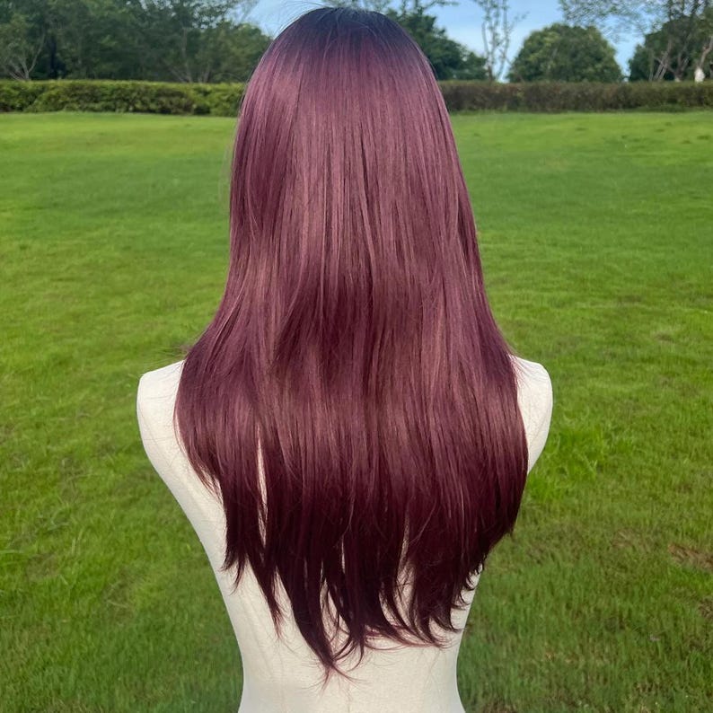 Op de afbeelding: Een lange, steile pruik in een donker bordeauxrode kleur, gepresenteerd op een mannequin. De pruik heeft een natuurlijke haarlijn en een gladde, strakke textuur. De achtergrond is een groene grasvlakte.