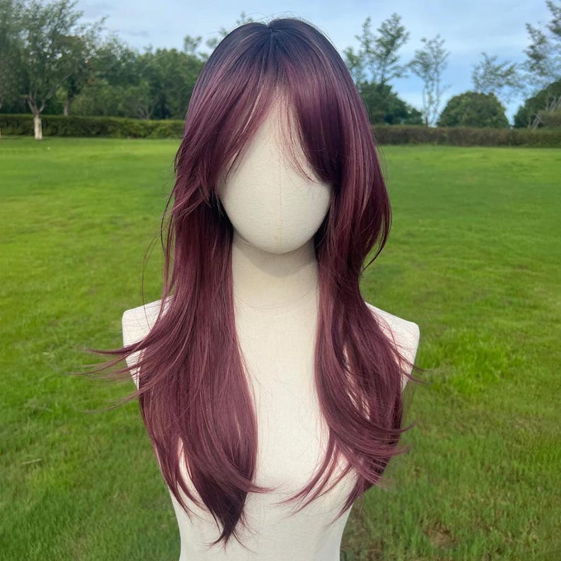 Op de afbeelding: Een lange, gelaagde pruik met een donker bordeauxrode kleur en een subtiel ombre-effect. De pruik heeft een zijwaarts geveegde pony en is gestyled op een mannequinhoofd. De achtergrond is een grasveld met bomen.