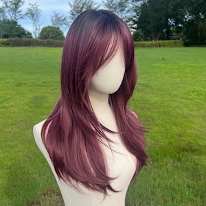 Op de afbeelding: Een lange, gelaagde pruik met donkere wortels en een diepe bordeauxrode tint. De pruik is gestyled met een zijwaartse pony en valt tot op de schouders. De pruik wordt getoond op een mannequinhoofd tegen een groene grasachtergrond.