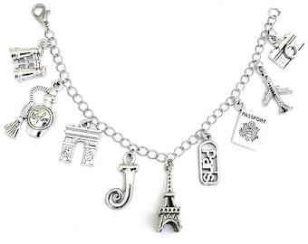 Könnte beinhalten: Silbernes Charm-Armband mit Reise-Themen-Anhängern. Zu den Anhängern gehören ein Miniatur-Eiffelturm, Arc de Triomphe, Flugzeug, Kamera, Reisepass, Parfümflasche, Fernglas und der Buchstabe "J". Das Armband ist mit einer Kette verbunden.
