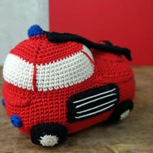 May include: A handmade crochet fire truck toy in red, white, black, and blue. The toy features a red body with white stripes, black wheels, and blue accents. The crochet work is detailed, showcasing the texture of the yarn.