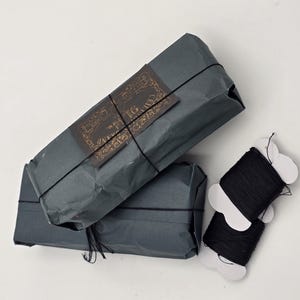 May include: Two packages wrapped in dark gray paper with black string and a label that says "The Mr. & Mrs. Collection". Two spools of black thread are wrapped in white paper with a cut-out design.