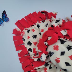 May include: A fleece blanket with a playing card design in red, black, and white. The blanket features a repeating pattern of hearts, spades, clubs, and diamonds. The edges are trimmed with red fringe. A blue and purple butterfly is in the upper left corner.