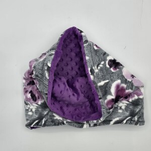 Op de afbeelding: Een grijze en paarse bloemenpatroon cowl sjaal met een diep paarse, getextureerde binnenkant. De sjaal is gemaakt van zachte stof en is ontworpen om om de nek te worden gedragen voor warmte en stijl.
