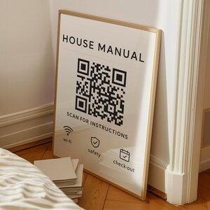 Puede incluir: Un póster blanco enmarcado con las palabras "HOUSE MANUAL" en negro, un gran código QR e iconos de wi-fi, seguridad y check-out. El marco es dorado y el póster está apoyado contra una pared sobre un suelo de madera.
