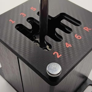 May include: A black gear shift assembly with a carbon fiber pattern top. The shift pattern is visible with red numbers and a chrome shift lever. Silver knobs are on the corners of the assembly.