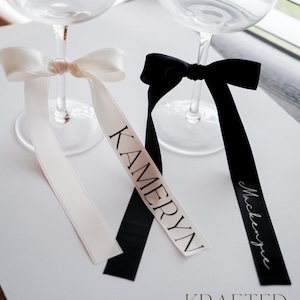 May include: Two champagne flutes with personalized ribbons tied around the stems. The white ribbon says "Kamerryn" and the black ribbon says "Mackenzie".